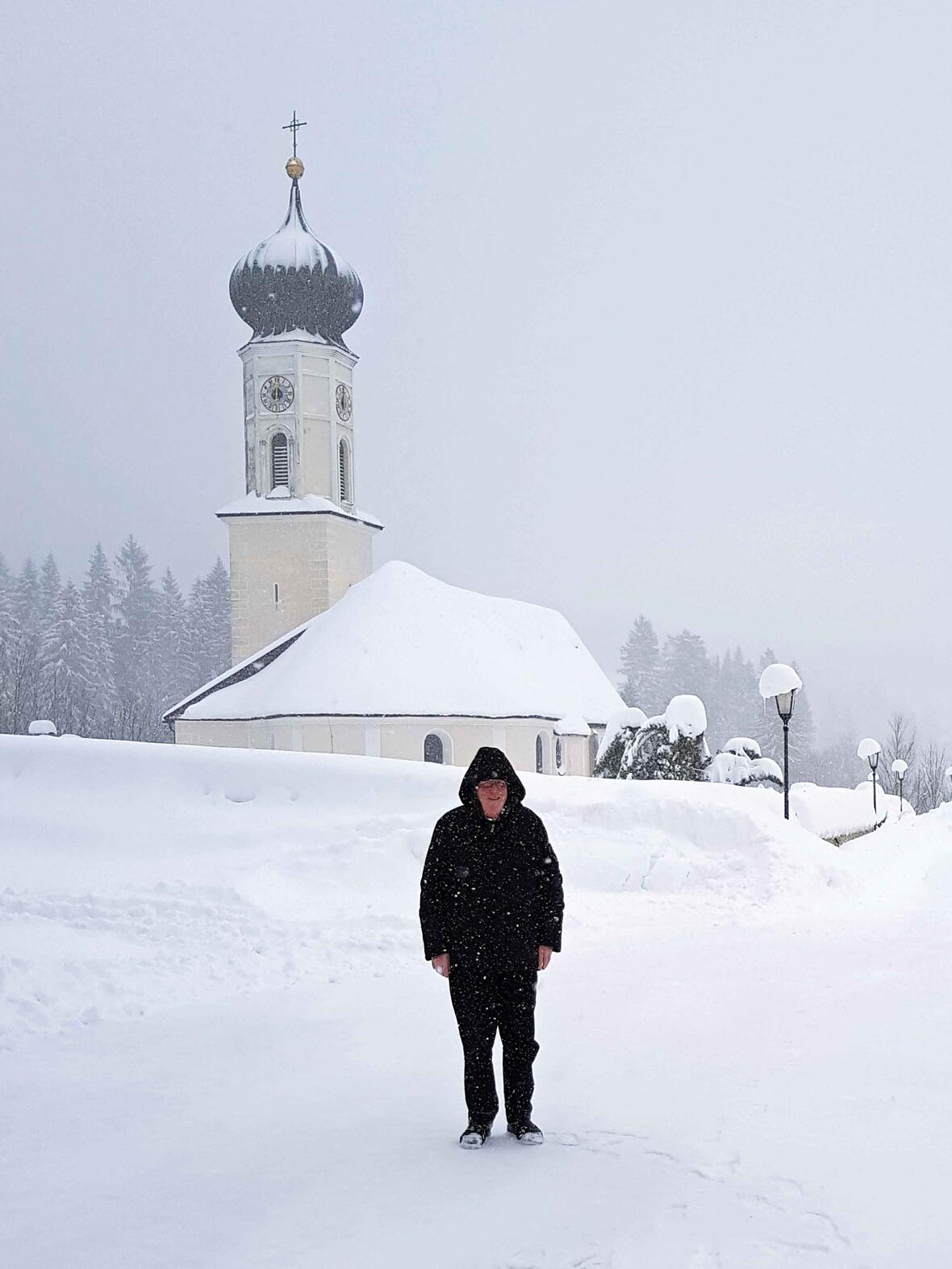 Nimmt die Situation mit Humor und einer gesunden Portion Gelassenheit: Pfarrer Willi Milz vor seiner von Schneemassen umgebenen Pfarrkirche St. Nikolaus. (Foto: privat)