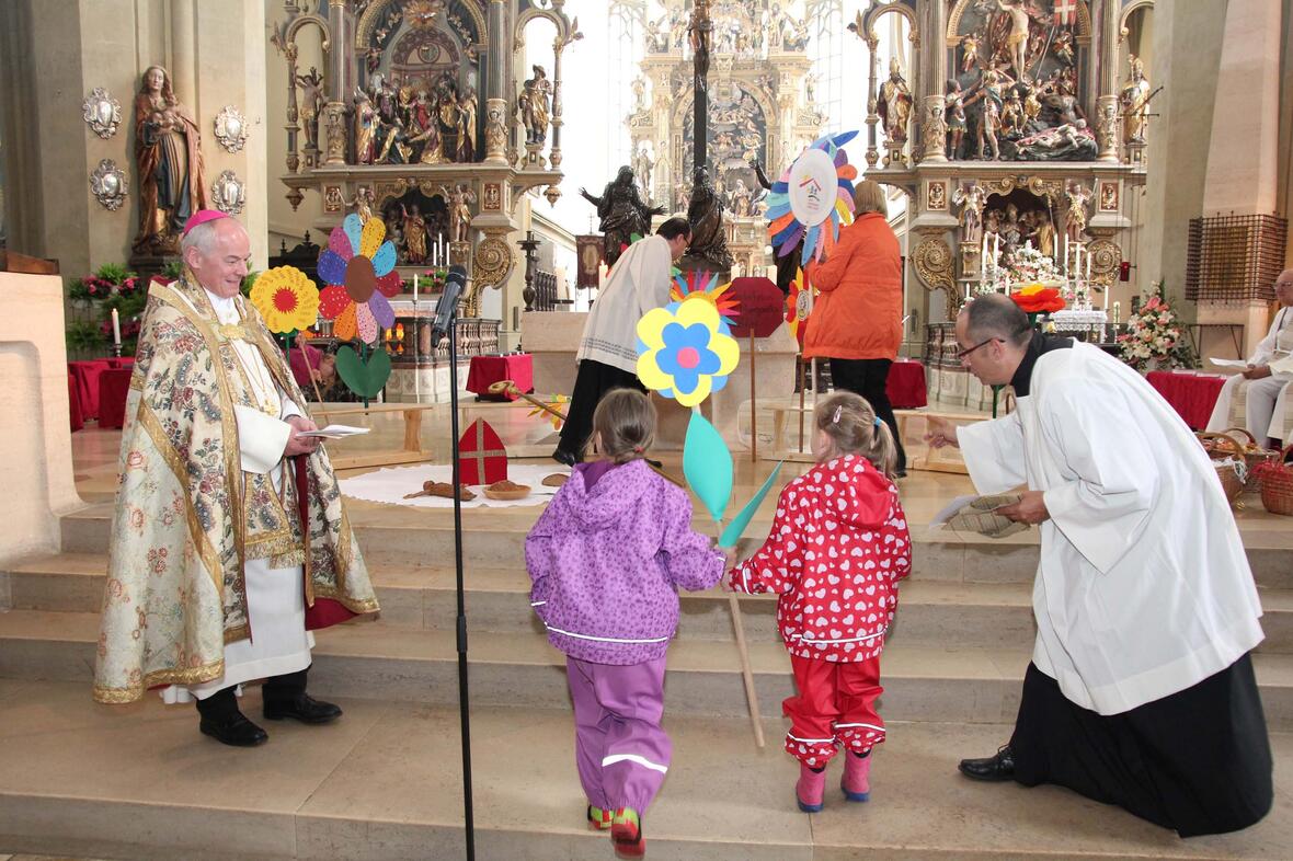 Kinder bringen ihre gebastelten Blumen als Gastgeschenk ihrer Kindertageseinrichtungen zum Altar. Weihbischof Losinger (l.) hatte ihnen erzählt, wie sehr sich der Bischof Ulrich um die armen und hungrigen Menschen gesorgt hatte. (Foto: Caritas)