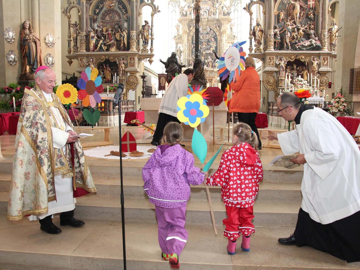 Kinder bringen ihre gebastelten Blumen als Gastgeschenk ihrer Kindertageseinrichtungen zum Altar. Weihbischof Losinger (l.) hatte ihnen erzählt, wie sehr sich der Bischof Ulrich um die armen und hungrigen Menschen gesorgt hatte. (Foto: Caritas)