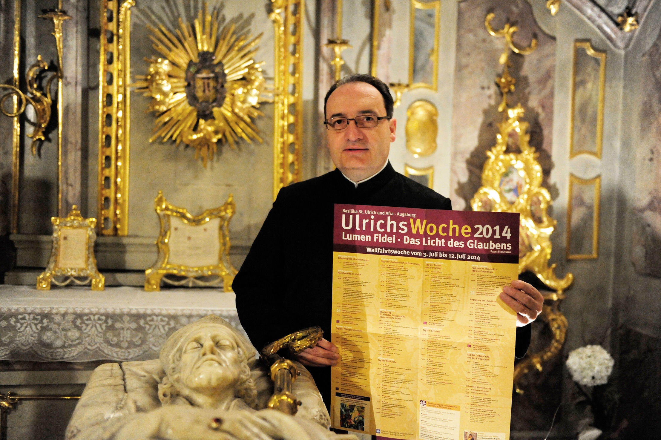 Stadtpfarrer Lothar Hartmann mit dem Plakat zur Ulrichswoche am Grab des hl. Ulrich (Bild: pba/Nicolas Schnall).