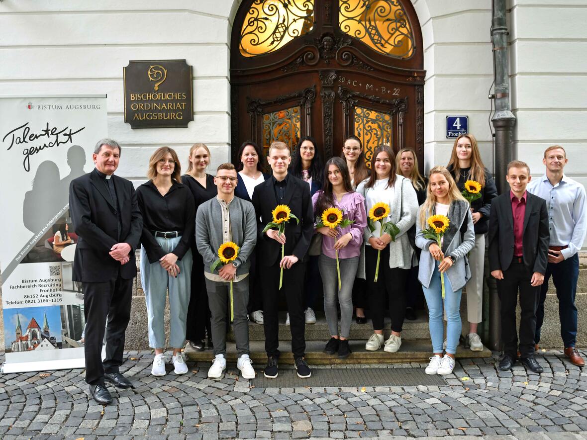 Herzlich mit Sonnenblumen begrüßt: Die neuen Auszubildenden zusammen mit Domkapitular Walter Schmiedel (Erster Stellvertretender Generalvikar), Vertretern der Personalabteilungen und die Jugendvertretung vor dem Bischöflichen Ordinariat. (Foto: Nicolas Schnall / pba). 